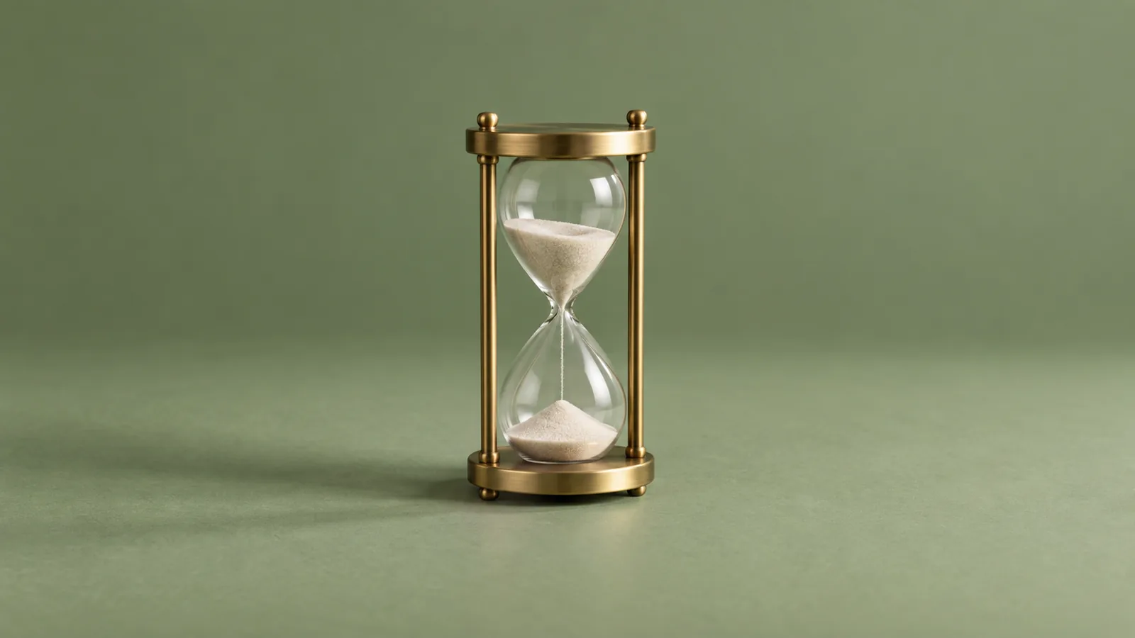 A small brass hourglass with pale sand falling through its center stands alone on a clean sage-green surface under soft side light.